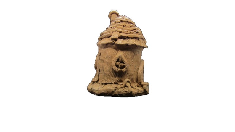 May include: A small, brown, clay-like fairy house with a thatched roof and a small window. The house is sitting on a base that looks like dirt.