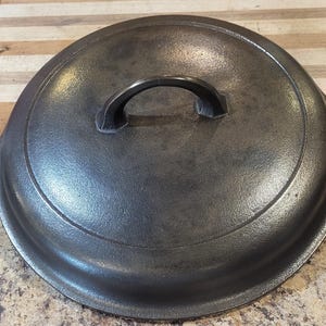 May include: A round, black cast iron lid featuring a central arched handle and a stepped rim design. The surface has a textured, matte finish with visible signs of use, including subtle variations in the dark metal tone across the top.
