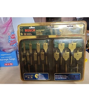 May include: A set of Bosch Rapidfeed spade bits in a clear plastic case. The bits are gold-colored and come in various sizes, with the largest bit labeled as 1 1/2 inches. The case includes the text "BONUS Folding Case".