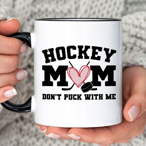 May include: White ceramic mug with a black rim and handle. The mug has a black and red graphic that says "Hockey Mom" with a heart and hockey sticks. The text below the graphic says "Don't Puck With Me".