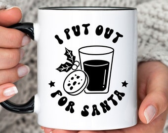 May include: White ceramic mug with a black rim and handle. The mug has a black and white graphic that says "I put out for Santa" with a glass of red liquid and a cookie.