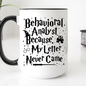 May include: White ceramic mug with a black handle and rim. The mug features the text "Behavioral Analyst Because My Letter Never Came" in black, along with whimsical illustrations of a wizard hat, castle, glasses, and a wand.