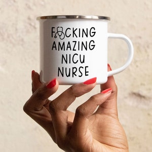Op de afbeelding: Een witte emaille mok met een zwart handvat en de tekst "F*CKING AMAZING NICU NURSE" met een stethoscoopicoon.