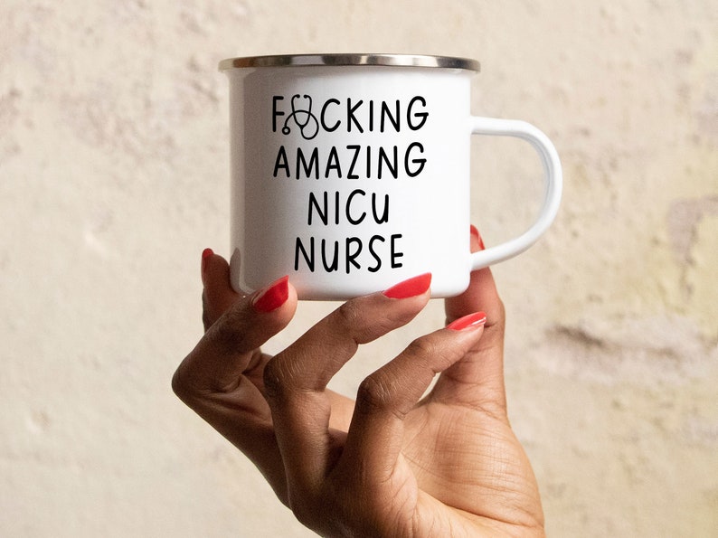 Op de afbeelding: Een witte emaille mok met een zwart handvat en de tekst "F*CKING AMAZING NICU NURSE" met een stethoscoopicoon.