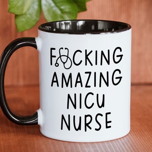 Op de afbeelding: Een witte koffiemok met een zwarte handgreep en een zwarte rand. De mok heeft zwarte tekst die "F*CKING AMAZING NICU NURSE" luidt met een stethoscoopsymbool.