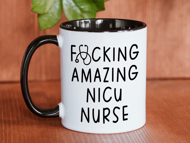 Op de afbeelding: Een witte koffiemok met een zwarte handgreep en een zwarte rand. De mok heeft zwarte tekst die "F*CKING AMAZING NICU NURSE" luidt met een stethoscoopsymbool.