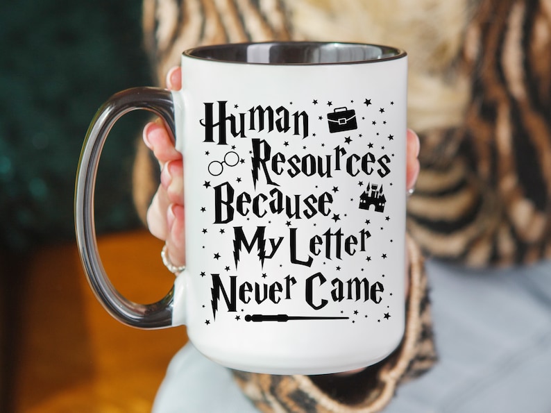 May include: White mug with a black rim and handle. The mug has a black graphic that says "Human Resources Because My Letter Never Came" with a Hogwarts castle, glasses, and a briefcase.