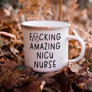 Op de afbeelding: Een witte emaille mok met een zwarte afbeelding waarop "F*CKING AMAZING NICU NURSE" staat met een stethoscoopsymbool. De mok staat op een bed van bruine bladeren.