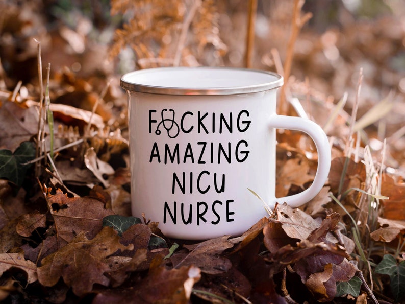 Op de afbeelding: Een witte emaille mok met een zwarte afbeelding waarop "F*CKING AMAZING NICU NURSE" staat met een stethoscoopsymbool. De mok staat op een bed van bruine bladeren.