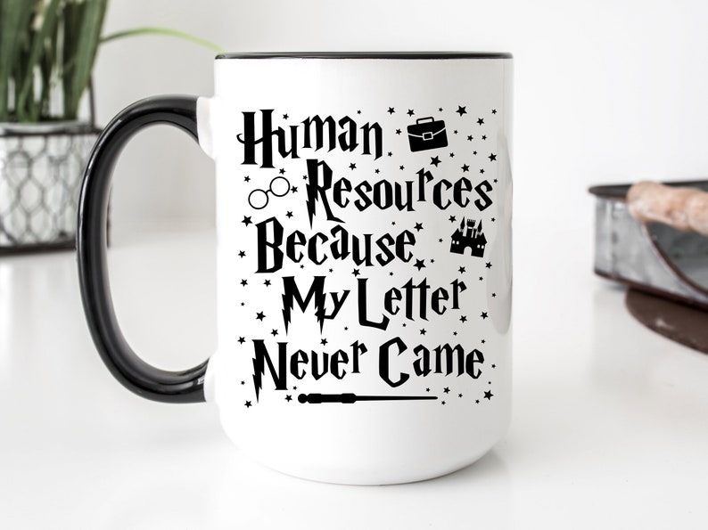 May include: White ceramic mug with a black handle and rim. The mug has a black graphic that says "Human Resources Because My Letter Never Came" with a wand, glasses, briefcase, and castle.