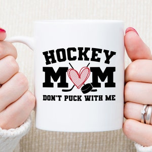 May include: White ceramic mug with a black and red design. The mug says "Hockey Mom" with a heart and hockey sticks. The text below says "Don't Puck With Me".