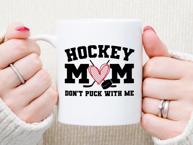 May include: White ceramic mug with a black and red design. The mug says "Hockey Mom" with a heart and hockey sticks. The text below says "Don't Puck With Me".
