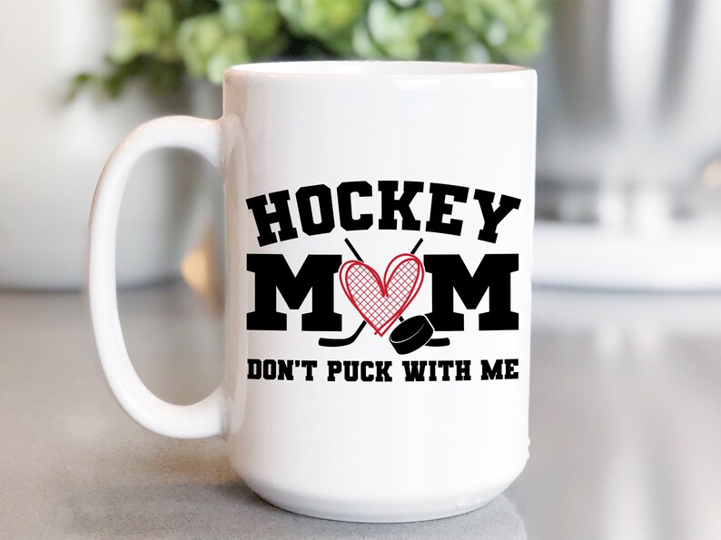 May include: White ceramic coffee mug with a black and red design. The mug features the text "HOCKEY MOM DON'T PUCK WITH ME" with a heart and hockey sticks.