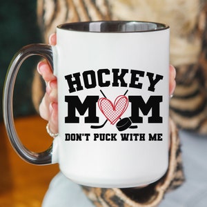 May include: White ceramic coffee mug with a black rim and handle. The mug has a black and red graphic that says "Hockey Mom" with a heart and hockey sticks. The text below the graphic says "Don't Puck With Me".