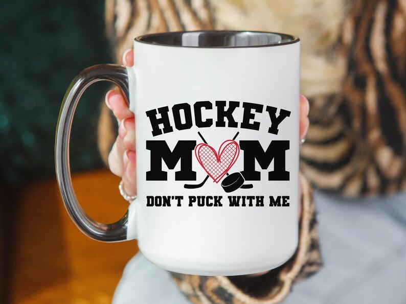 May include: White ceramic coffee mug with a black rim and handle. The mug has a black and red graphic that says "Hockey Mom" with a heart and hockey sticks. The text below the graphic says "Don't Puck With Me".
