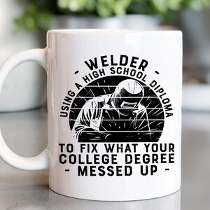 May include: White ceramic coffee mug with a black graphic of a welder wearing a welding mask and the text "Welder - Using a High School Diploma to fix what your college degree messed up - "