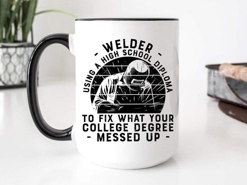 May include: White ceramic coffee mug with a black handle and black rim. The mug features a black and white illustration of a welder with the text "USING A HIGH SCHOOL DIPLOMA TO FIX WHAT YOUR COLLEGE DEGREE MESSED UP - WELDER - "