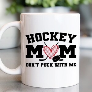May include: White ceramic coffee mug with a black graphic that says "Hockey Mom" with a red heart and hockey sticks. The text below the graphic says "Don't Puck With Me".