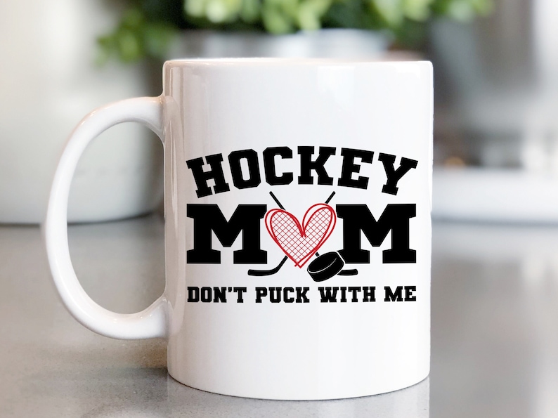 May include: White ceramic coffee mug with a black graphic that says "Hockey Mom" with a red heart and hockey sticks. The text below the graphic says "Don't Puck With Me".