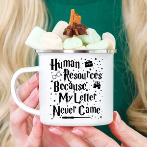May include: White enamel mug with a black design that says "Human Resources Because My Letter Never Came" with a graphic of a briefcase, glasses, a castle, and a wand. The mug is filled with marshmallows and topped with cinnamon sticks and a star anise.