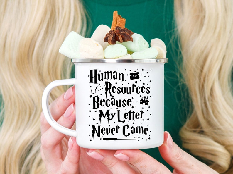 May include: White enamel mug with a black design that says "Human Resources Because My Letter Never Came" with a graphic of a briefcase, glasses, a castle, and a wand. The mug is filled with marshmallows and topped with cinnamon sticks and a star anise.