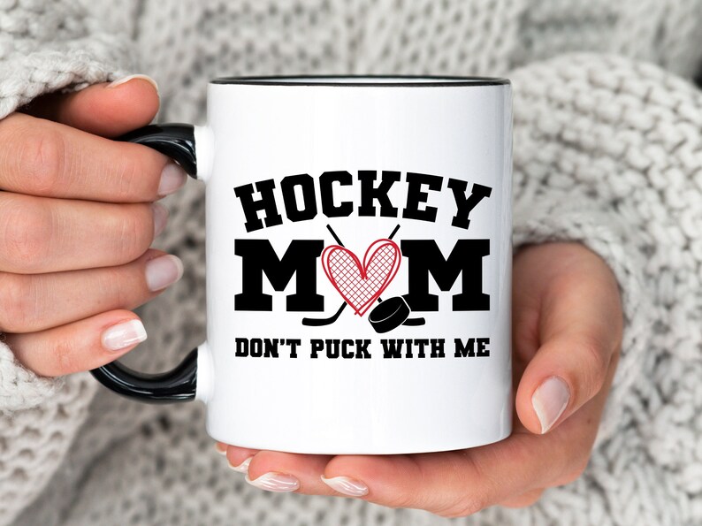 May include: White ceramic mug with a black rim and handle. The mug has a black and red graphic that says "Hockey Mom" with a heart and hockey sticks. The text below the graphic says "Don't Puck With Me".