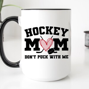 May include: White ceramic coffee mug with a black handle and rim. The mug has a black graphic that says "Hockey Mom" with a red heart and hockey sticks. The text below the graphic says "Don't Puck With Me".