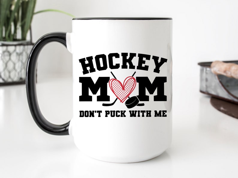 May include: White ceramic coffee mug with a black handle and rim. The mug has a black graphic that says "Hockey Mom" with a red heart and hockey sticks. The text below the graphic says "Don't Puck With Me".