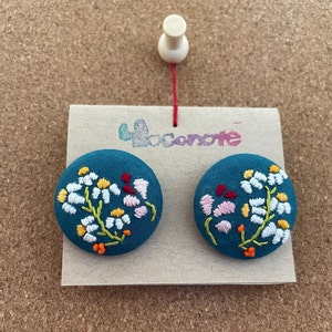 以下が含まれることがあります： 白、ピンク、黄色、オレンジの花が刺繍された、ティールブルーの布製のボタン2つ。ボタンは、「locanote」と印刷されたカードに表示されています。