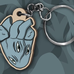 May include: A wooden keychain shaped like a blue anatomical heart with three eyes. The keychain has a silver metal ring.