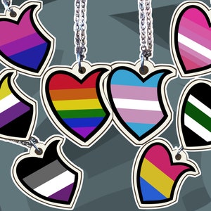 Op de afbeelding: Zeven hartvormige hangers aan zilveren kettingen, elk met een ander ontwerp van de pride-vlag: biseksueel, regenboog, transgender, non-binair, genderqueer, lesbisch en panseksueel.