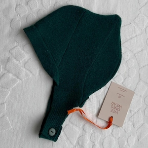 May include: A dark green baby bonnet with a button closure and an attached tag. The bonnet is made of a soft, textured fabric. The tag is attached with an orange ribbon and features the text "WEE ONES WEAR".
