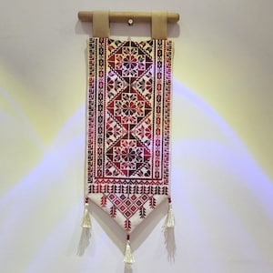 May include: A white tapestry with a red and white geometric pattern, hanging on a white wall. The tapestry has tassels on the bottom.