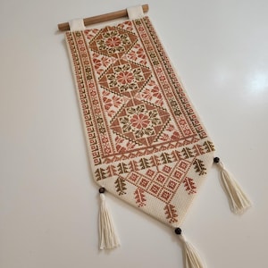 Puede incluir: Un tapiz de pared rectangular blanquecino con un diseño detallado de punto de cruz en tonos marrón, rojo y verde. Presenta patrones geométricos y florales, una barra de madera en la parte superior y borlas en la parte inferior. Mide unos 61 cm de largo.