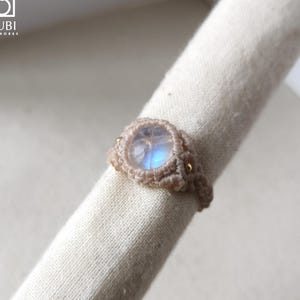 Peut inclure: Une bague en macramé faite à la main avec une pierre précieuse ronde et irisée. La bague est fabriquée avec du cordon de macramé de couleur beige et ornée de petites perles dorées. La bague est présentée sur un tissu blanc.