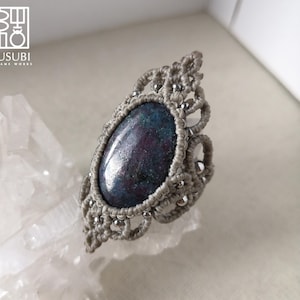 Puede incluir: Un anillo de macramé gris con una piedra preciosa azul y morada. El anillo está hecho con cuentas plateadas y tiene un diseño delicado e intrincado.