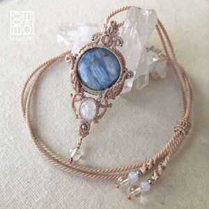 Puede incluir: Un collar de macramé hecho a mano con una piedra azul redonda grande y una piedra blanca ovalada más pequeña. El colgante está intrincadamente tejido con un cordón beige y adornado con pequeñas cuentas. El collar tiene un cordón largo y ajustable.