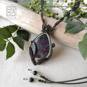 Può includere: Una collana in macramè con un grande ciondolo in pietra a forma di goccia, di colore rosso scuro e nero, incastonato in un cordino annodato marrone scuro. Il cordino include piccole perline verdi e trasparenti, con due estremità decorate.