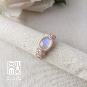 Op de afbeelding: Handgemaakte macramé ring met een ovale maansteen, met blauwe en witte tinten. De ring is gemaakt met lichtroze macramé koord en kleine zilveren kralen. De ring wordt gepresenteerd op een witte doek. Het logo "MUSUBI" is zichtbaar.