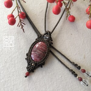 May include: A red and white striped stone pendant necklace with a brown macrame cord. The pendant is oval shaped and has a clear crystal bead above it. The necklace has two brown cords with red and clear beads at the ends.