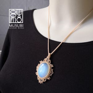 Puede incluir: Un colgante de ópalo azul claro con un diseño de macramé, colgado de un collar de cordón crema.