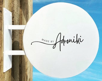 May include: White round sign with black text that reads "Made by Adroniki" against a blue sky background.