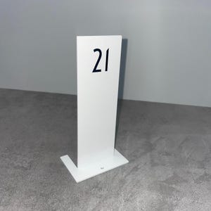 Può includere: Un segnaposto bianco con il numero "21" stampato in nero. Il segnaposto ha una forma rettangolare con una base. Lo sfondo è un muro grigio chiaro e un pavimento grigio.