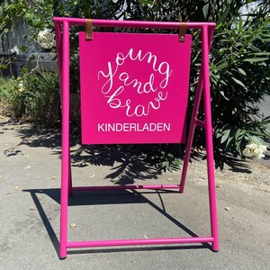 May include: A pink A-frame sign with the words "young and brave" written in white cursive lettering. The sign also says "kinderladen" in white lettering.