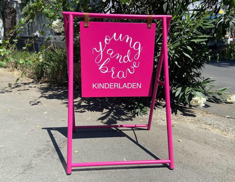 May include: A pink A-frame sign with the words "young and brave" written in white cursive lettering. The sign also says "kinderladen" in white lettering.