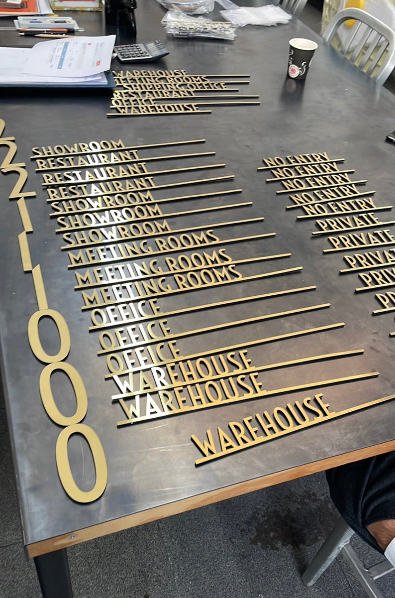 May include: A close-up of gold-colored metal signs with various room names and numbers. The signs are arranged in rows on a dark gray surface. The signs include: Showroom, Restaurant, Meeting Rooms, Office, and Warehouse.