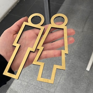 May include: Two gold-colored metal cutout figures of a person, one with a skirt and one with pants. The figures are on a light-colored surface.