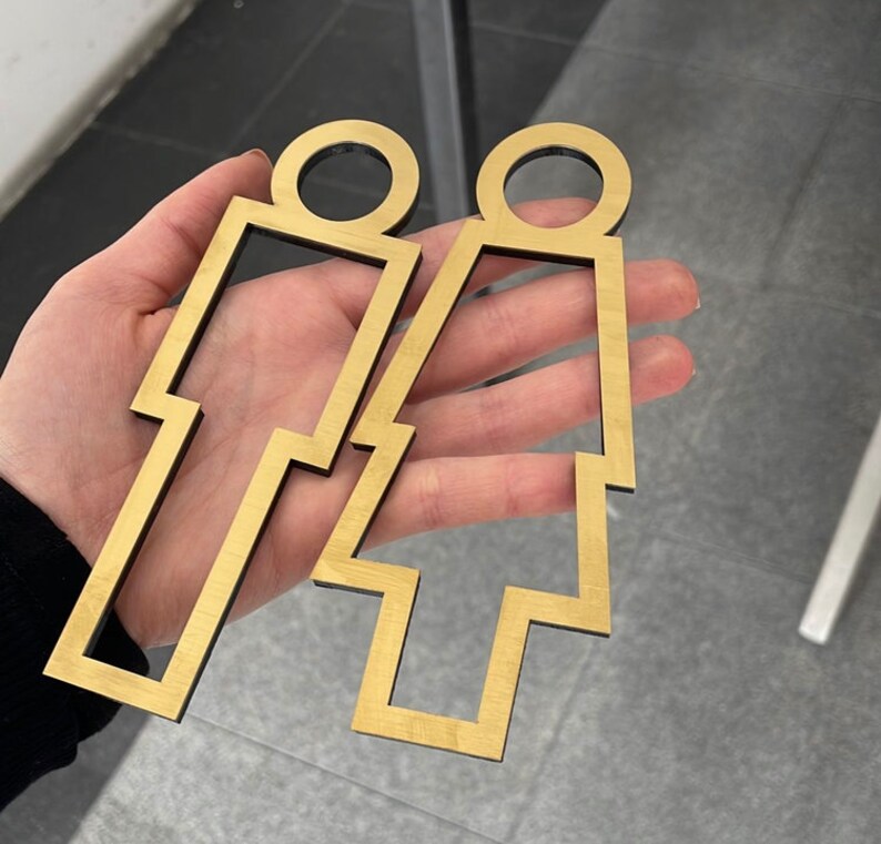 May include: Two gold-colored metal cutout figures of a person, one with a skirt and one with pants. The figures are on a light-colored surface.
