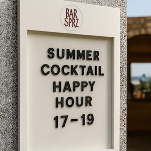 May include: A rectangular white sign with a raised frame featuring the text BAR SPRZ at the top, followed by the black lettering SUMMER COCKTAIL HAPPY HOUR 17-19. The sign is mounted on a textured gray stone wall, displaying clear, bold typography.