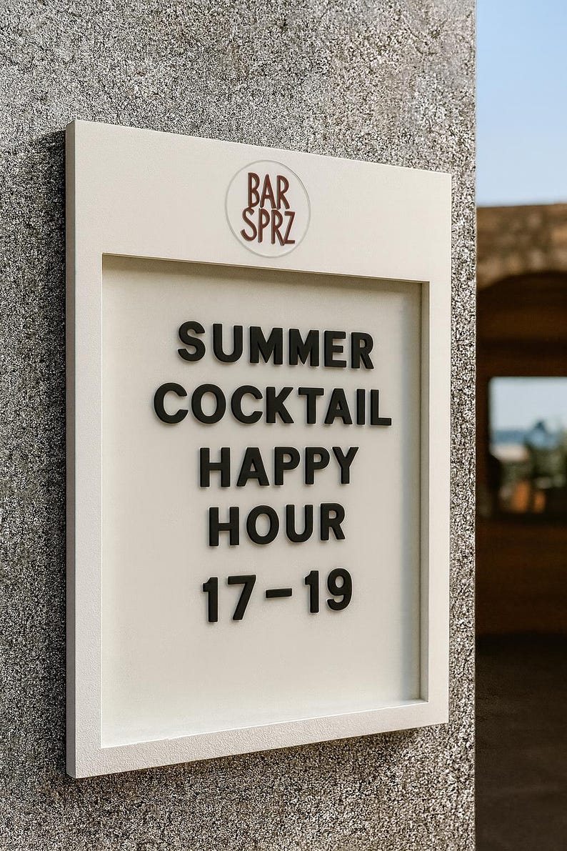 May include: A rectangular white sign with a raised frame featuring the text BAR SPRZ at the top, followed by the black lettering SUMMER COCKTAIL HAPPY HOUR 17-19. The sign is mounted on a textured gray stone wall, displaying clear, bold typography.